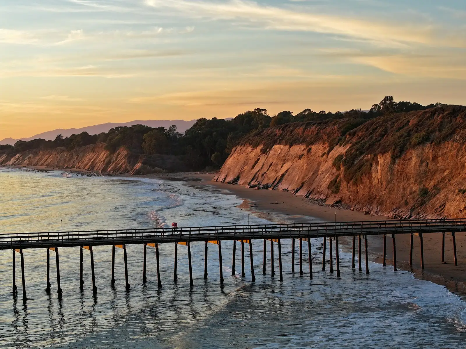 A wooden pier extending into the ocean beside colorful bluffs at sunset in Goleta, California.
